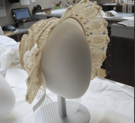 Front of a cream lace cap embroidered with raised gold swirling motifs and scalloped edging, mounted on a display stand in a studio workspace.